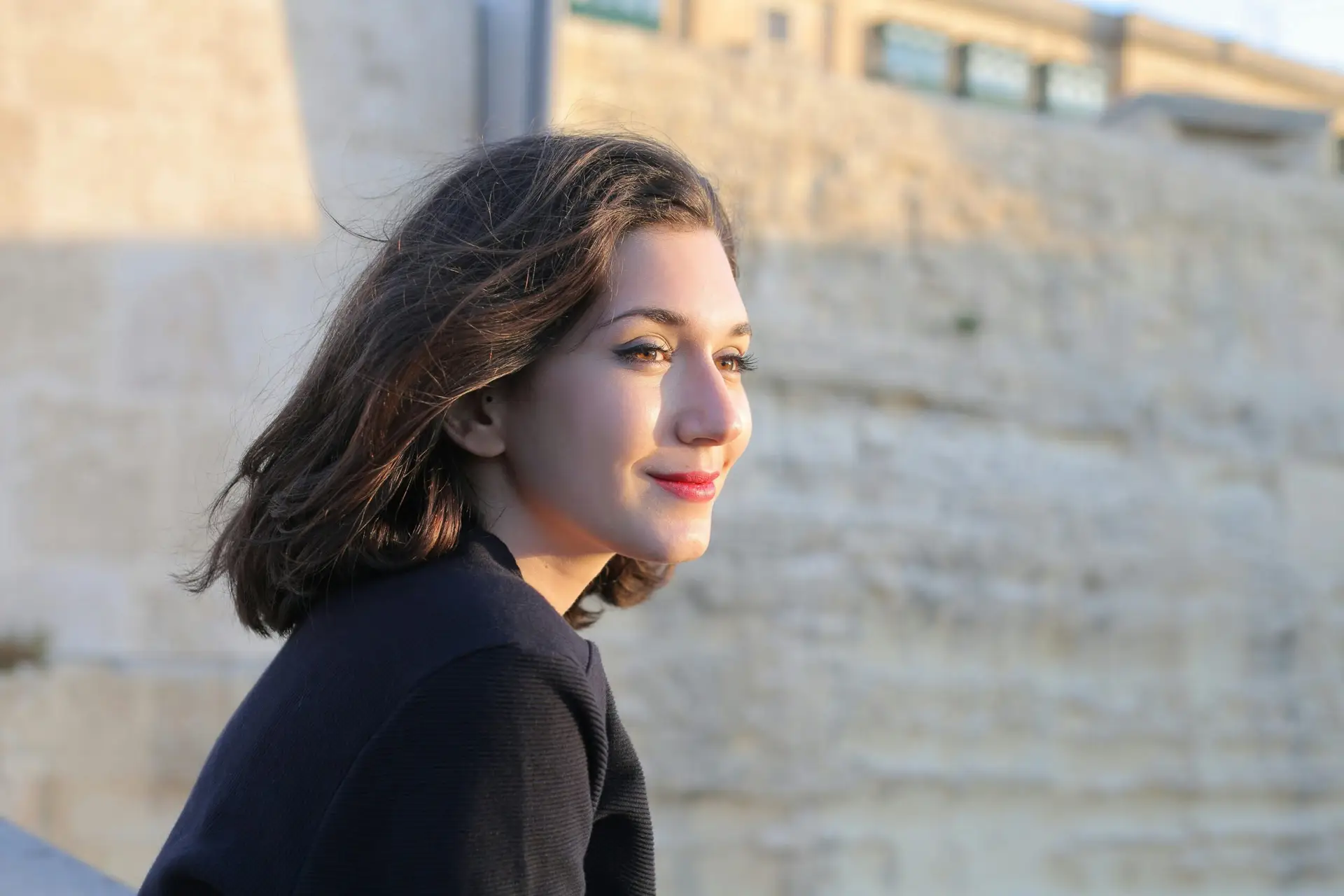 A young woman smiling outdoors near a historical stone wall in Mgarr, Malta.